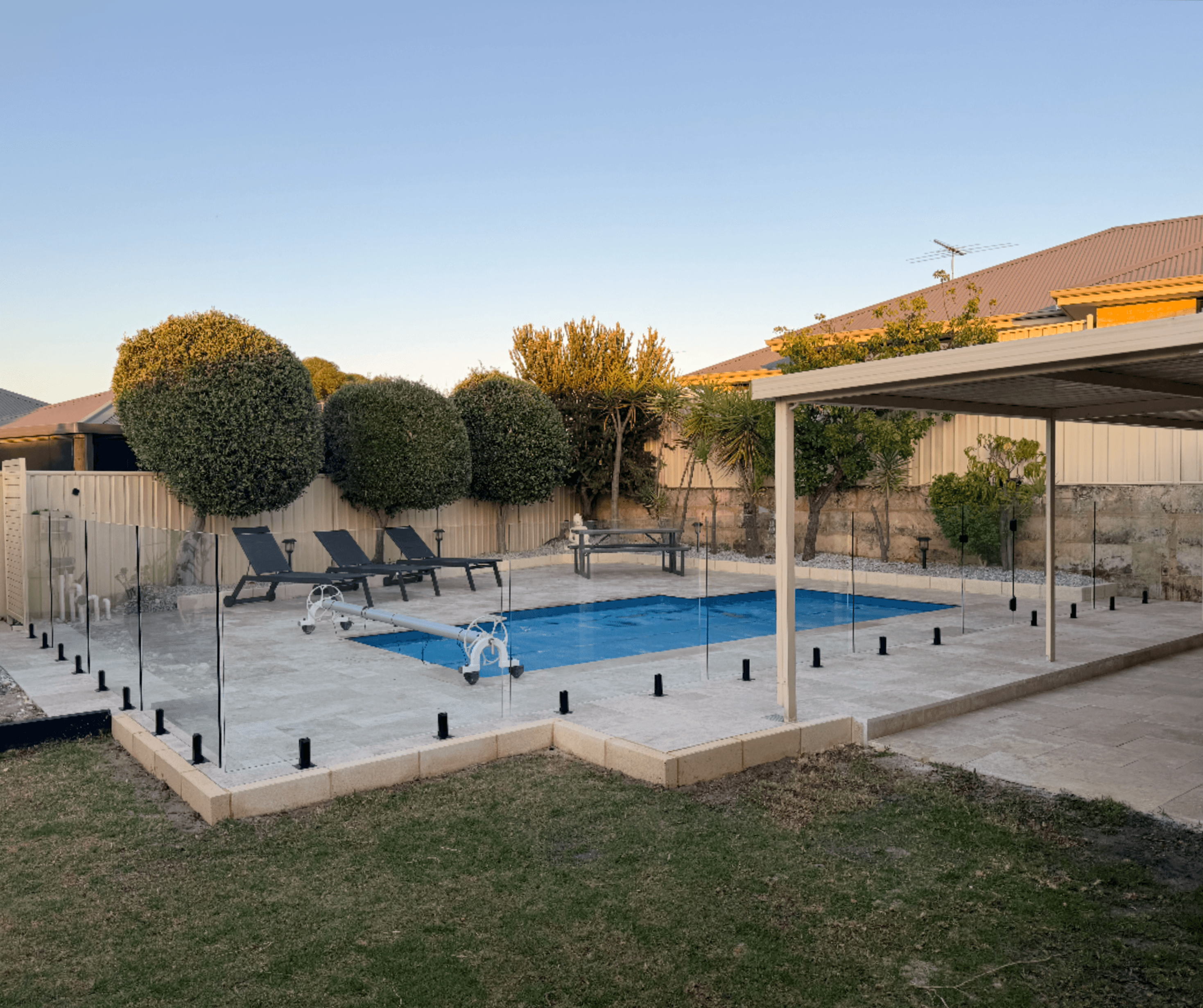 Paved pool surround behind frameless glass fencing, with clipped round trees at dusk
