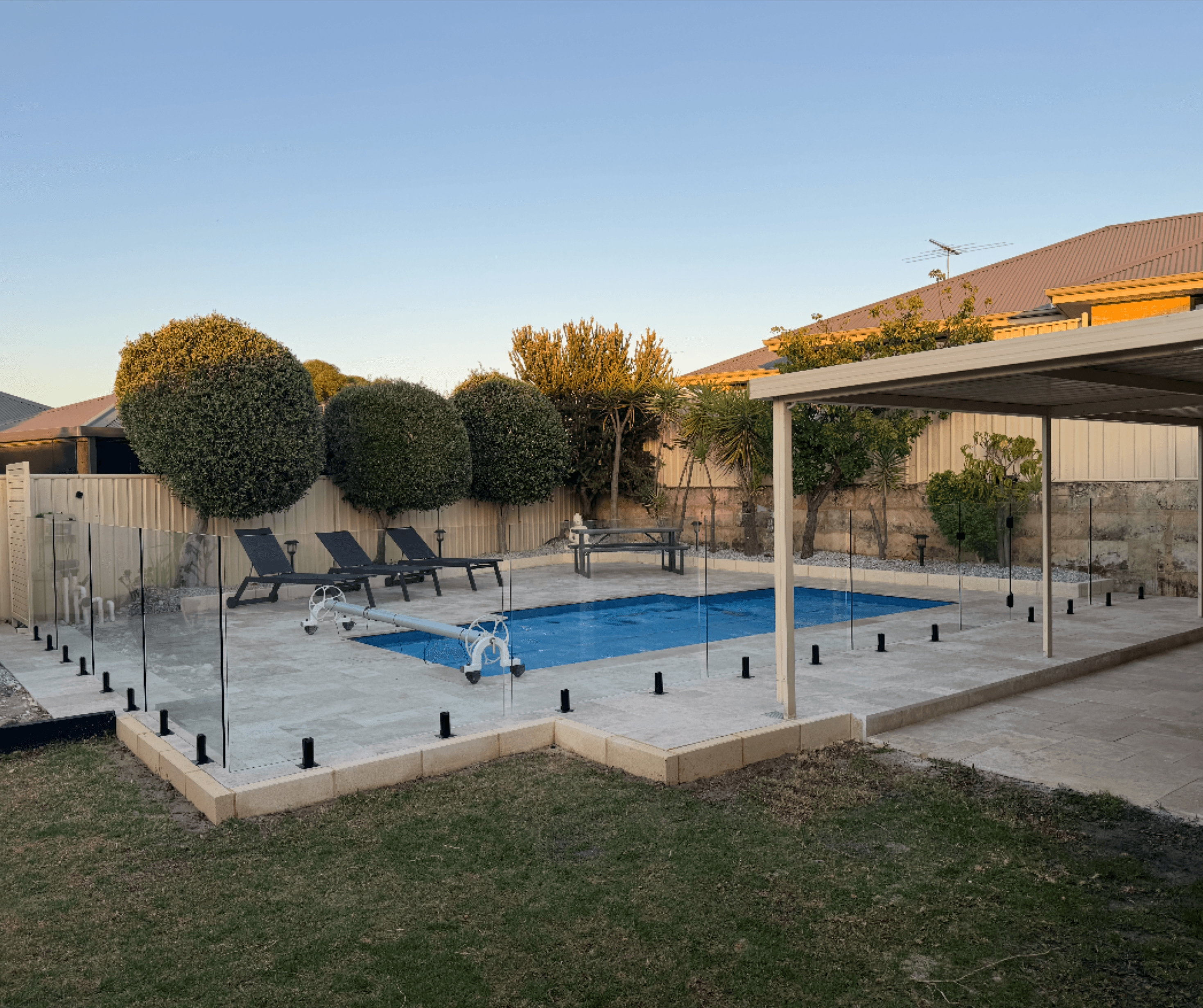 Paved pool surround behind frameless glass fencing, with clipped round trees at dusk